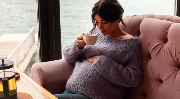 Pregnant woman in a purple sweater holding a cup of tea, resting her hand on her belly.