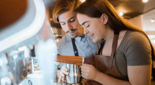 Två personer lär sig barista-yrket vid en espressomaskin. Hon håller en mjölkkanna, han tittar på.