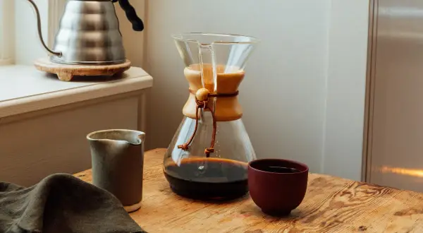 En Chemex med färdigbryggt dropp kaffe står på ett träbord. Bredvid finns en liten kanna och en kopp. En vattenkokare syns i bakgrunden.