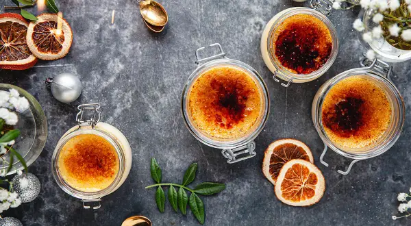 Fyra krämiga creme brulee i glasburkar, toppade med karamelliserat socker. En festlig dukning med torkad apelsin, julgranskulor och gnistrande stjärntändare.