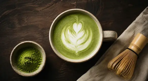 En kopp vackert dekorerad matcha latte med ett hjärtmönster, bredvid en skål med matchapulver och en bambuvisp.