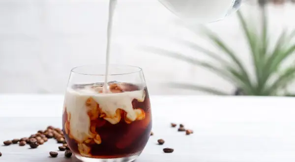 Mjölk hälls i ett glas med iskaffe på snabbkaffe. Kaffebönor ligger utspridda på bordet.