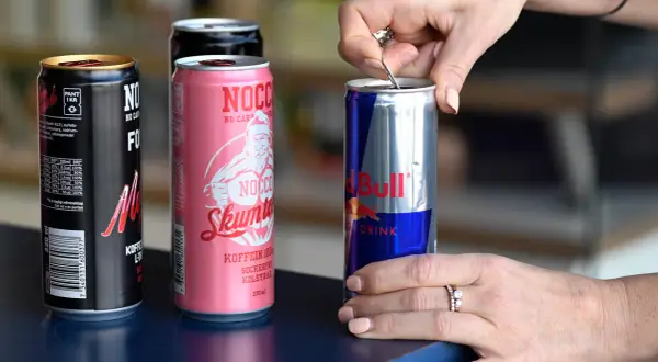 En hand öppnar en burk Red Bull, bredvid NOCCO-burkar. Koffein redbull för energi.