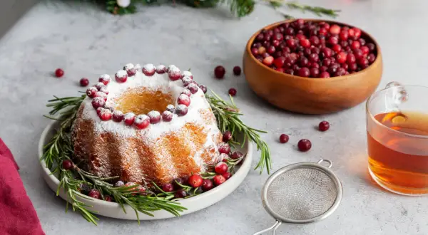 En vackert dekorerad sockerkaka med lingon och rosmarin, perfekt för julfika till många. En skål med lingon och en kopp te står redo.