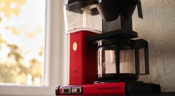 Röd Moccamaster kaffebryggare vid fönstret, redo att brygga en god kopp kaffe.