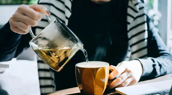 En person häller upp ett uppiggande te i en orange mugg. Ånga stiger från koppen.