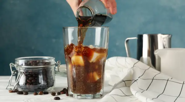 En hand häller mörkt kaffe över is i ett glas. Bredvid står en burk med kaffebönor. Kaffe smakar surt om det inte är perfekt bryggt.