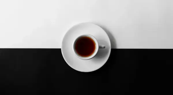 En vit kopp med mörkt kaffe står på en vit tefat, delar en yta som är uppdelad i svart och vitt. Kan kaffe bli gammalt?