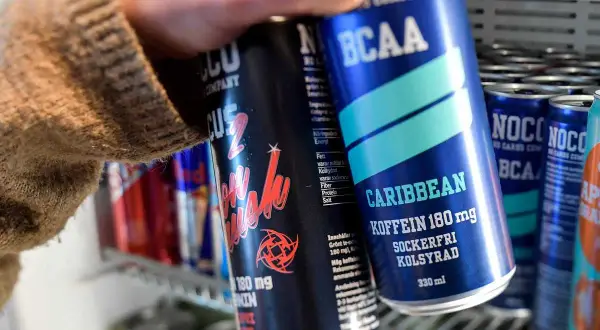 En hand tar en NOCCO Caribbean ur ett kylskåp fyllt med energidrycker. Frågan "hur många nocco per dag" dyker upp när man ser alla dessa.