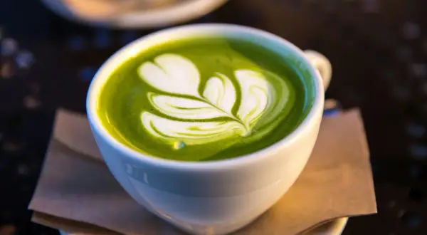 En vit kopp med grön matcha latte, dekorerad med ett vitt mönster som liknar ett löv. Matcha innehåll i sin finaste form.