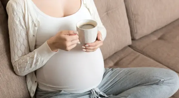 En gravid kvinna sitter på en soffa och håller en mugg med kaffe.