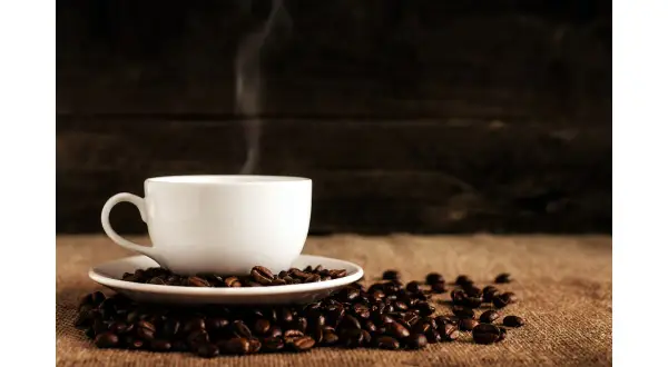 En vit kaffekopp i dl, omgiven av rostade kaffebönor, med ånga som stiger upp.