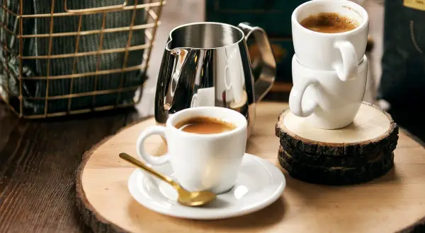 Enkel espresso i vit kopp med guldfärgad sked. En mjölkkanna och en stapel koppar står bredvid.
