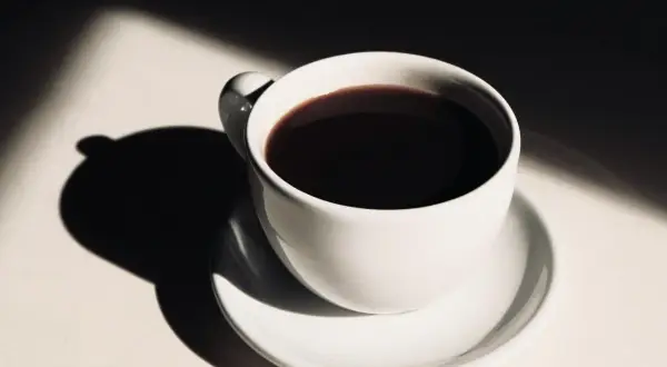 En vit kaffekopp med mörkt kaffe, belyst av solljus som skapar dramatiska skuggor. Hur påverkar kaffe blodtrycket?