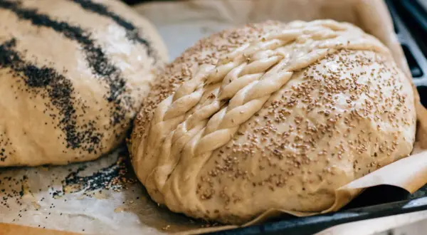 Två nybakade bröd på bakplåtspapper, ett toppat med vallmofrön och ett med sesamfrön och flätat mönster. Perfekta fika idéer!