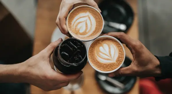 Tre händer håller varsin kopp med kaffe. Två koppar har vacker latte art, en är en kaffe macchiato.