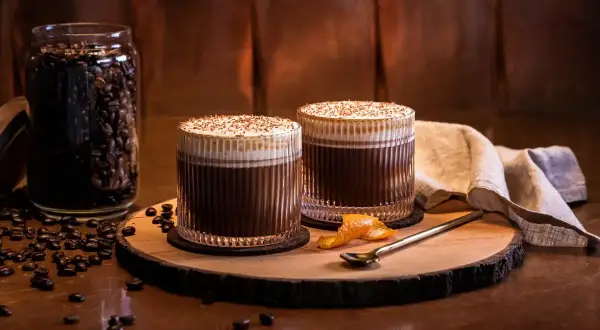 Två glas med krämig kaffe, toppad med vispad grädde och strössel. Bredvid står en burk med robusta kaffebönor.