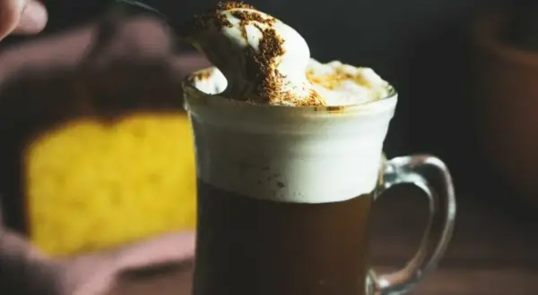 En sked med vispad grädde och kanel toppar en varm kaffe Karlsson utan Baileys. I bakgrunden syns en gul kaka.