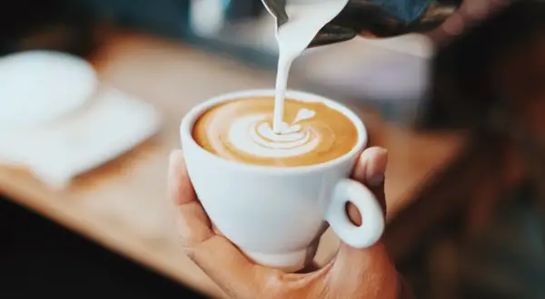 En hand håller en vit kopp med eritreansk kaffe, där mjölk hälls i för att skapa lattekonst.