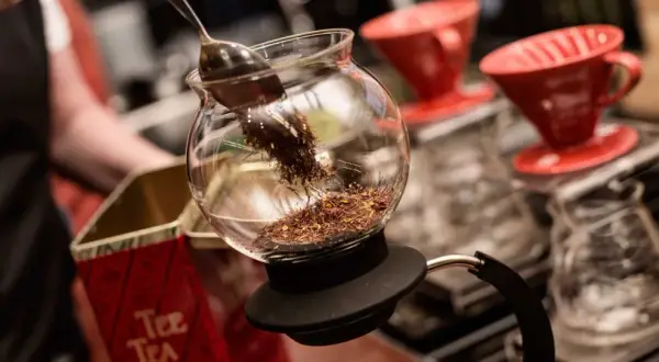 En sked lägger teblad i en glaskaraff, redo för att brygga te utan koffein. Röda kaffefilter står i bakgrunden.