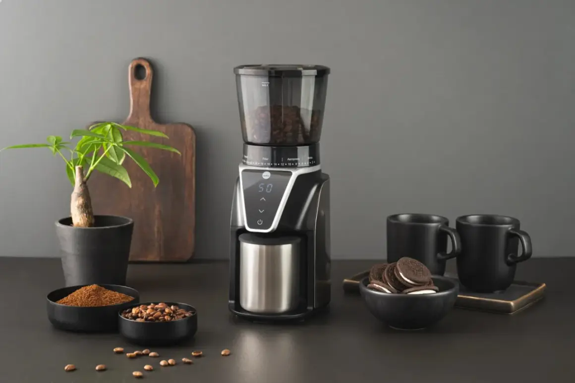 En modern kaffekvarn med digital display, fylld med bönor, bredvid skålar med malet kaffe och hela bönor. Två svarta muggar och en liten krukväxt fulländar scenen.