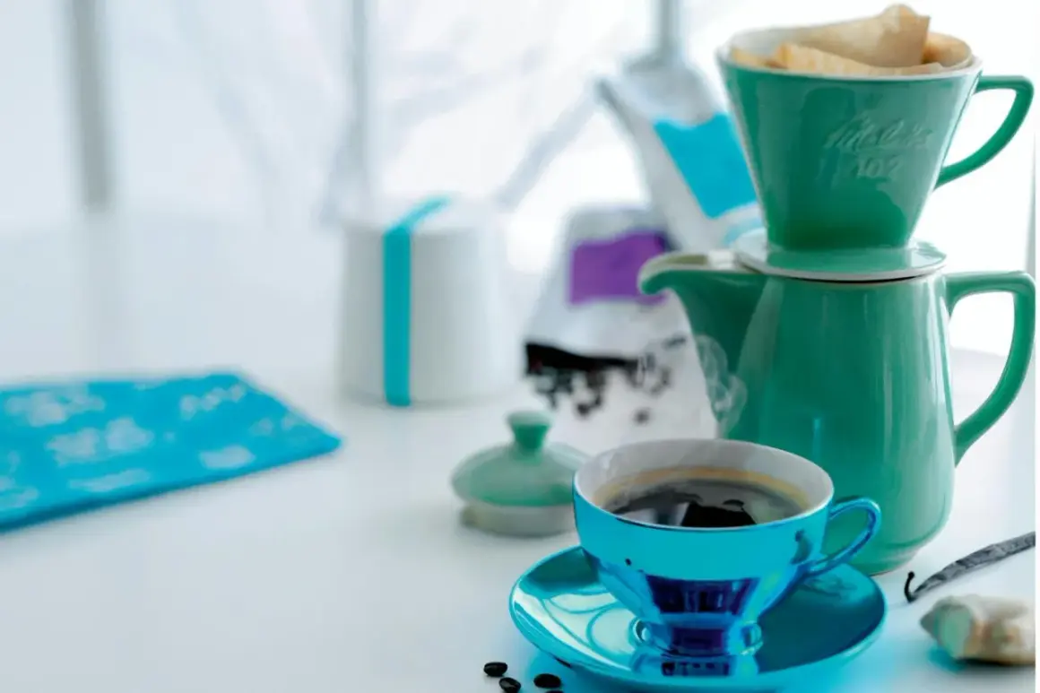 En turkos kaffekopp med fat står framför en grön kaffekanna med filterhållare. Kaffebönor och en vaniljstång ligger bredvid. En fin morgon med gott kaffe.