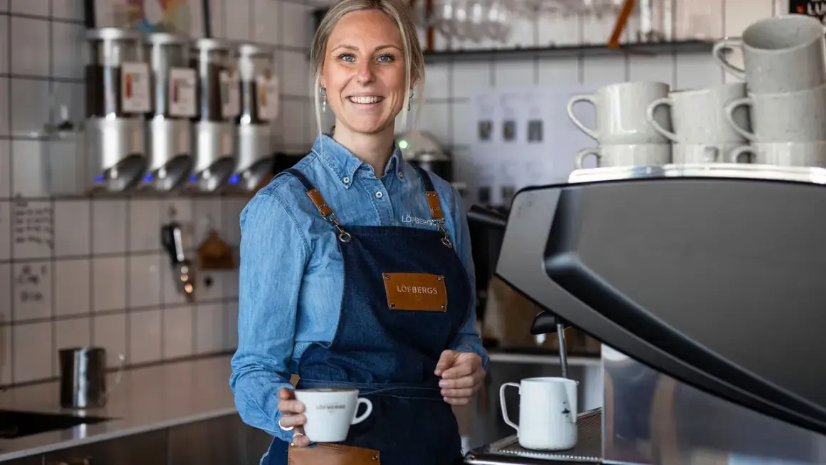 En leende barista håller en kaffekopp. Kanske en påminnelse om hur mycket kaffe dricker svenskar?