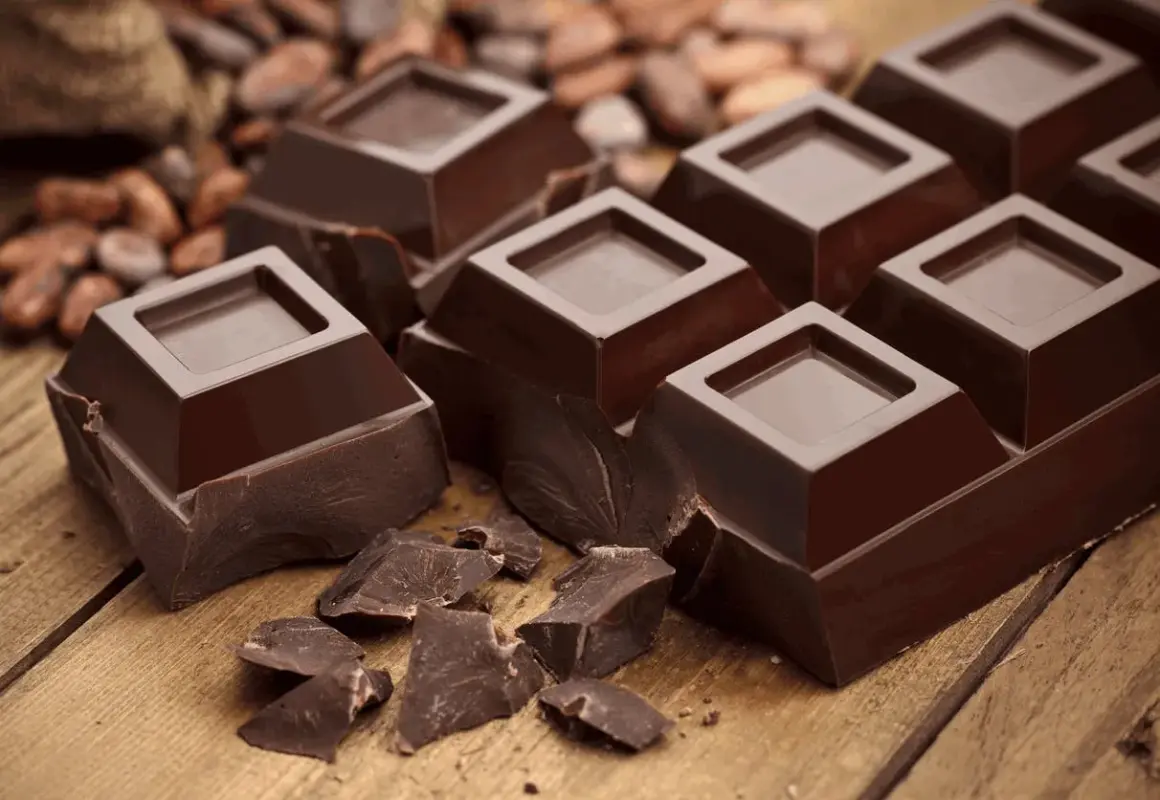 Mörk chokladbitar på träbord, med kakaobönor i bakgrunden. En härlig källa till kakao och koffein för en energikick.