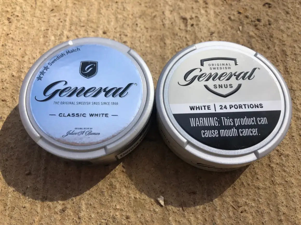 Två dosor General snus, en Classic White och en White, ligger på en sandig yta.