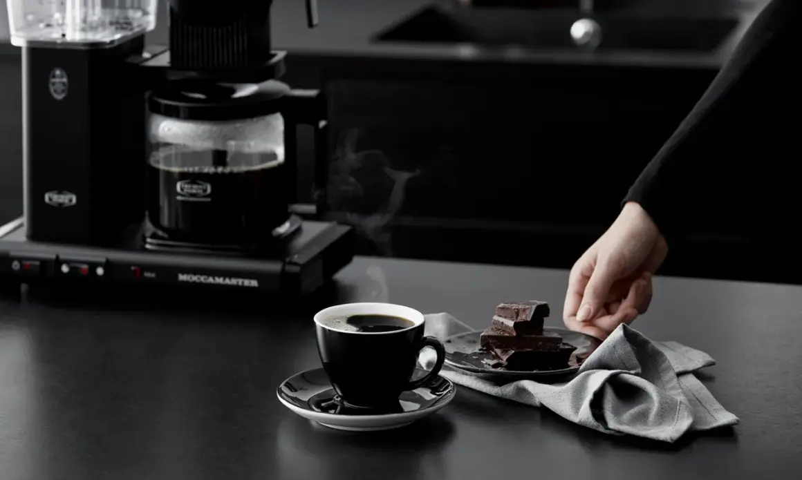 En hand sträcker sig efter chokladbitar bredvid en kopp ångande kaffe från en Moccamaster. Kanske är det dåligt kaffe, men chokladen ser god ut.