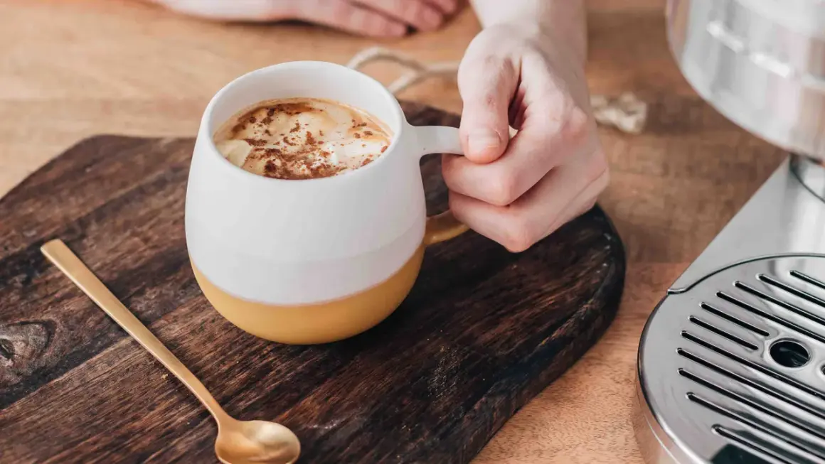 En hand håller en mugg med en krämig pumpa latte, toppad med kanel. En guldig sked ligger bredvid.