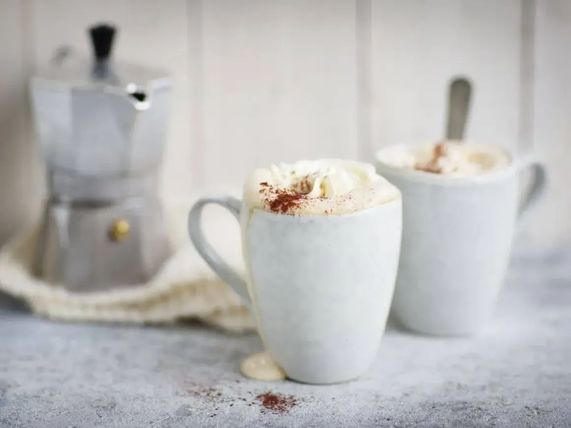 Två muggar med krämig mocca kaffe, toppad med vispgrädde och kakao. En mokabryggare står i bakgrunden, redo för ett nytt mocca kaffe recept.