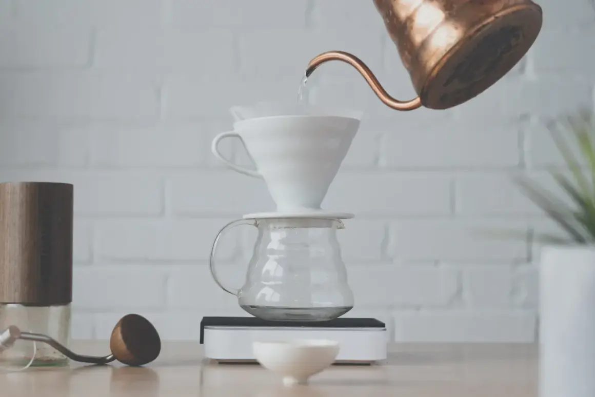 Vatten hälls från en kopparkanna över ett filter i en vit V60-bryggare, som står på en glaskaraff. En kaffekvarn och en skopa ligger bredvid. Detta visar hur brygger man kaffe.