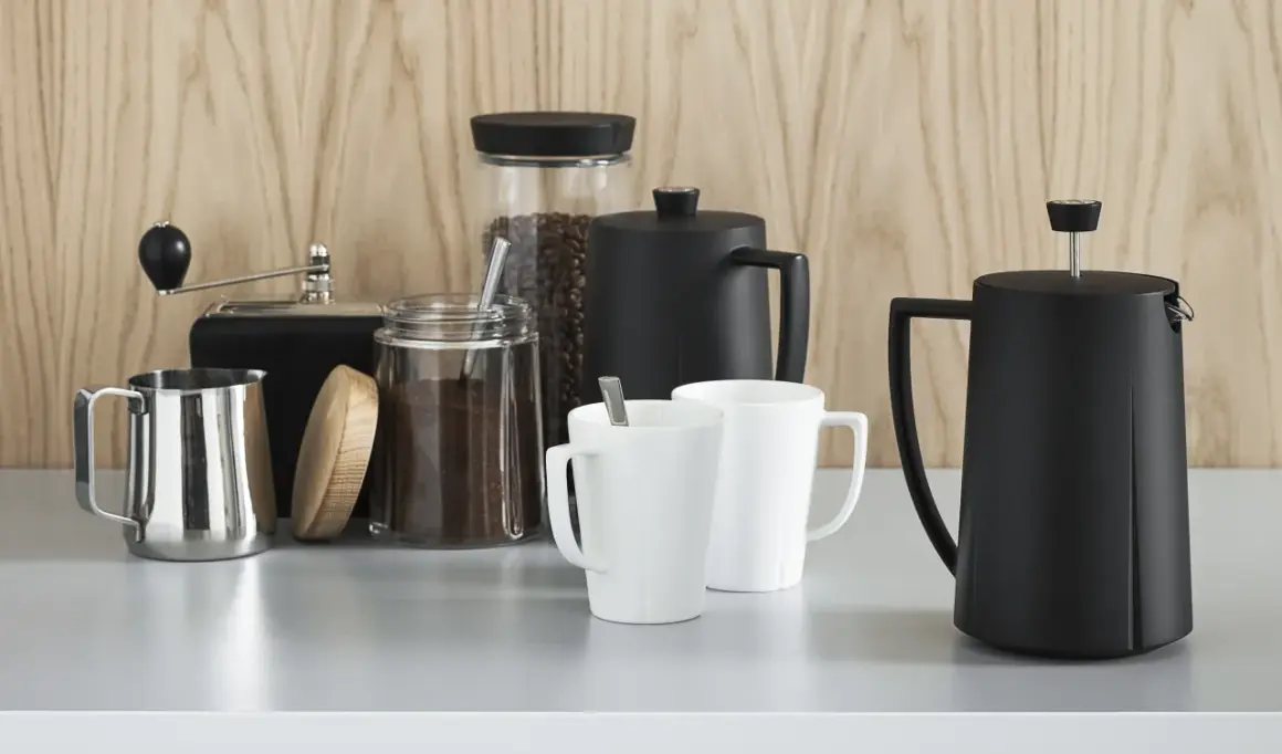 Kaffebönor och malet kaffe i glasburkar, en kaffekvarn, två vita muggar och två svarta presskannor. Perfekt för att utforska olika presskaffe sorter.