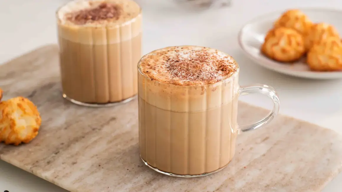 Två glas med krämigt skummande cafe americano, toppade med kanel. Bredvid står en liten tallrik med gyllene kokoskakor.