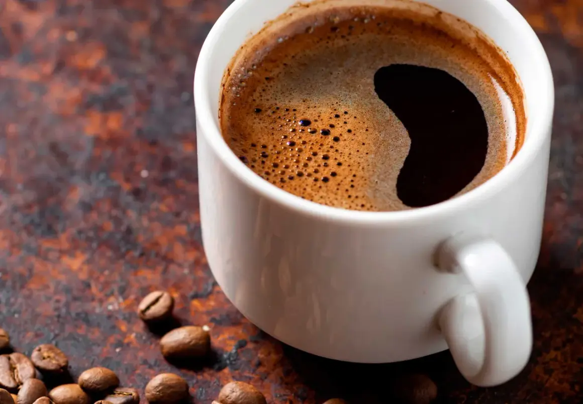 En vit kopp med starkt kaffe och skummig yta, omgiven av kaffebönor på en rustik bakgrund.