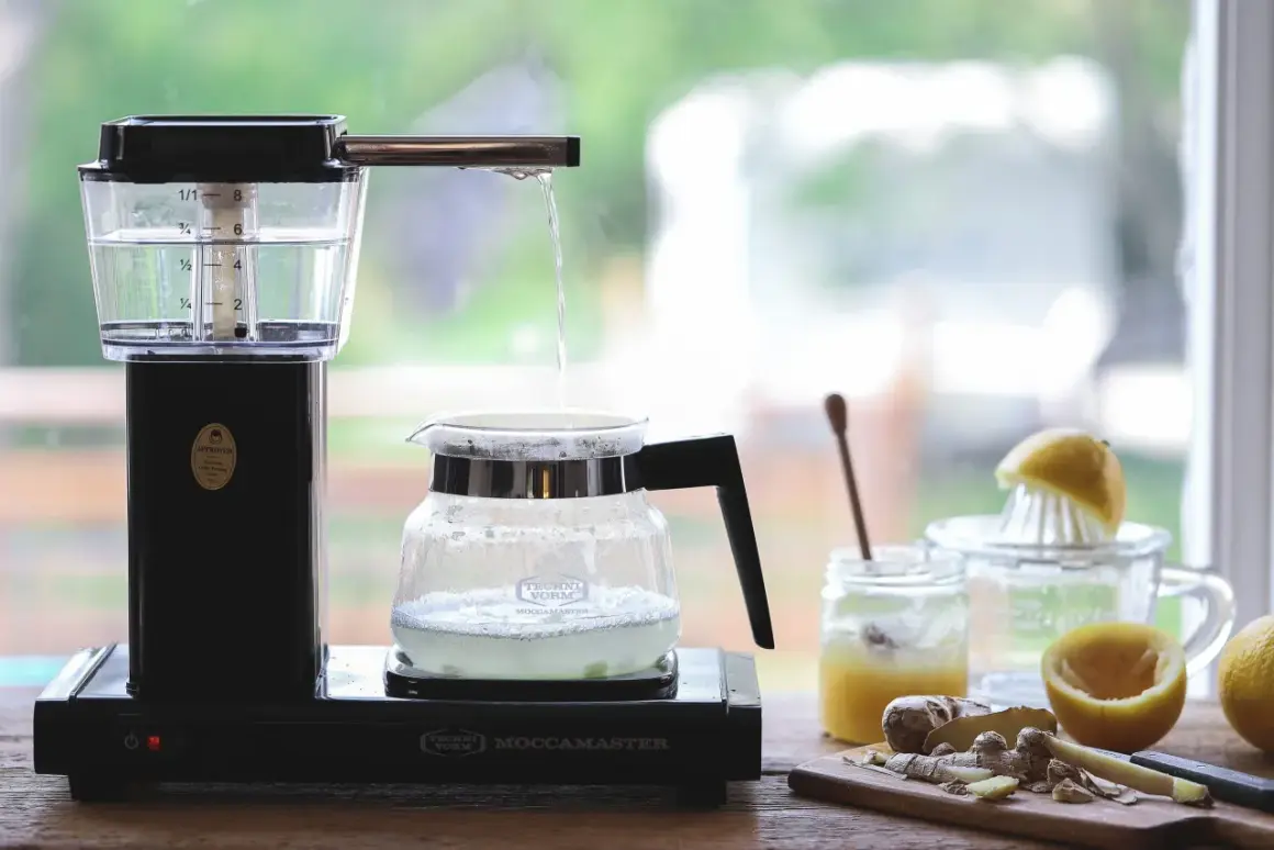 Vatten droppar från en kaffebryggare ner i en glaskanna, redo att brygga te. Ingredienser som citron och ingefära syns i bakgrunden.