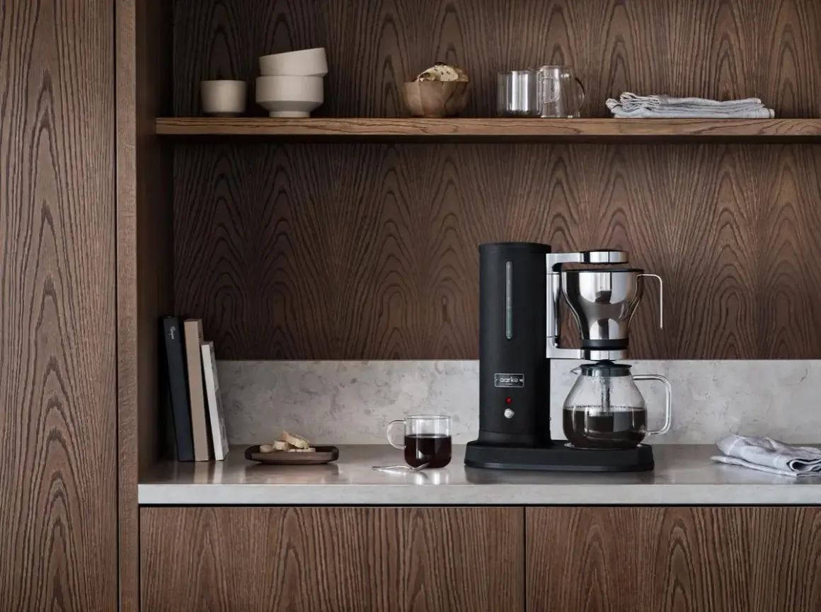 En stilren kaffebryggare i svart och krom, bredvid en glaskaraff med kaffe. På hyllan ovanför finns skålar och glas. En av flera olika kaffebryggare som passar in i ett modernt kök.