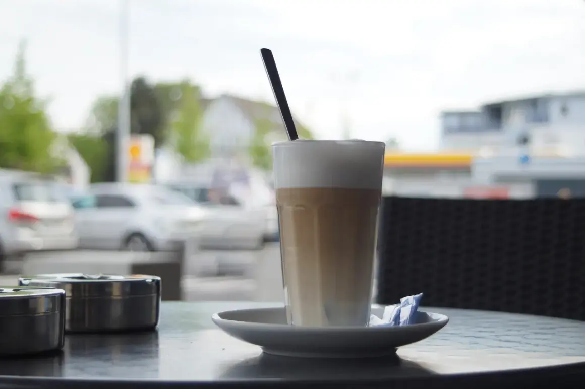 En hög latte macchiato med skummig mjölk och en sked, redo att avnjutas vid ett cafébord utomhus.