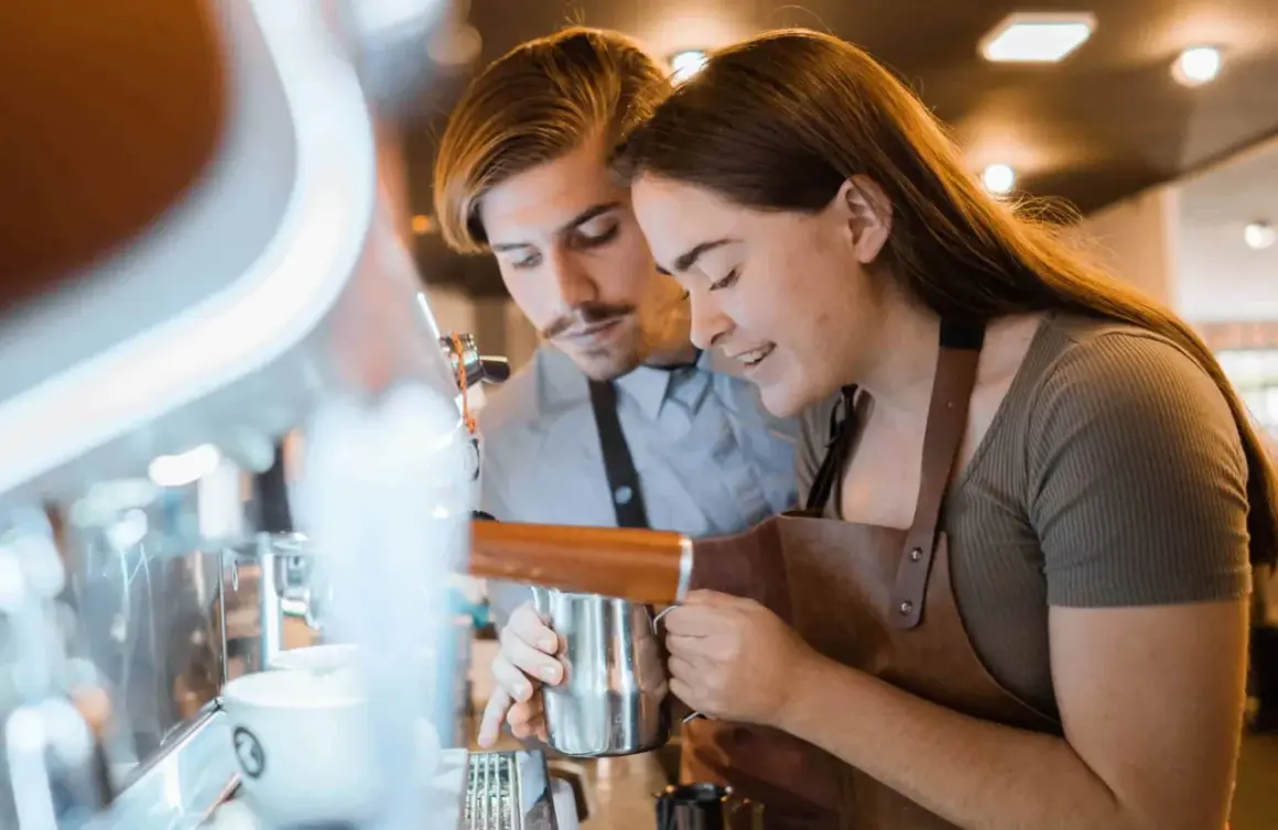 Två personer lär sig barista-yrket vid en espressomaskin. Hon håller en mjölkkanna, han tittar på.