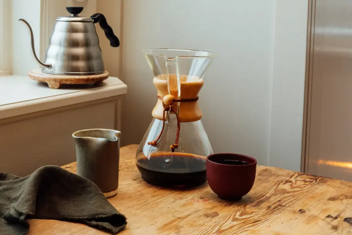 En Chemex med färdigbryggt dropp kaffe står på ett träbord. Bredvid finns en liten kanna och en kopp. En vattenkokare syns i bakgrunden.