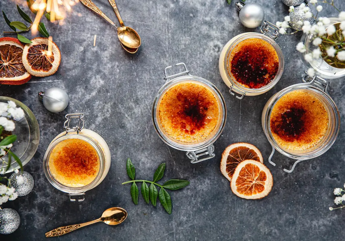 Fyra krämiga creme brulee i glasburkar, toppade med karamelliserat socker. En festlig dukning med torkad apelsin, julgranskulor och gnistrande stjärntändare.