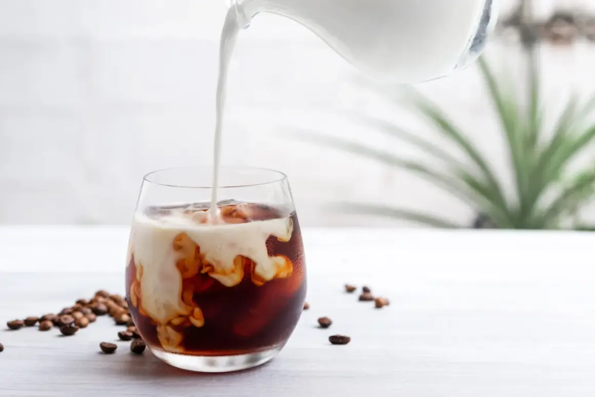 Mjölk hälls i ett glas med iskaffe på snabbkaffe. Kaffebönor ligger utspridda på bordet.