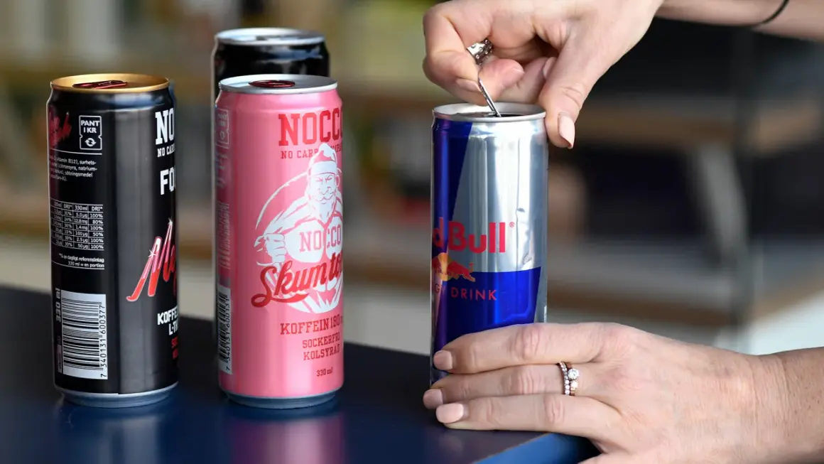 En hand öppnar en burk Red Bull, bredvid NOCCO-burkar. Koffein redbull för energi.