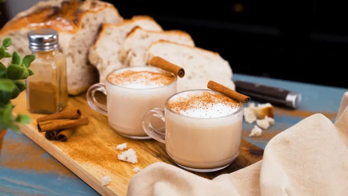 Två muggar krämig chai latte med kanelstänger och strödd kanel. Bröd och kryddor i bakgrunden. Perfekt för ett mysigt chai latte recept.