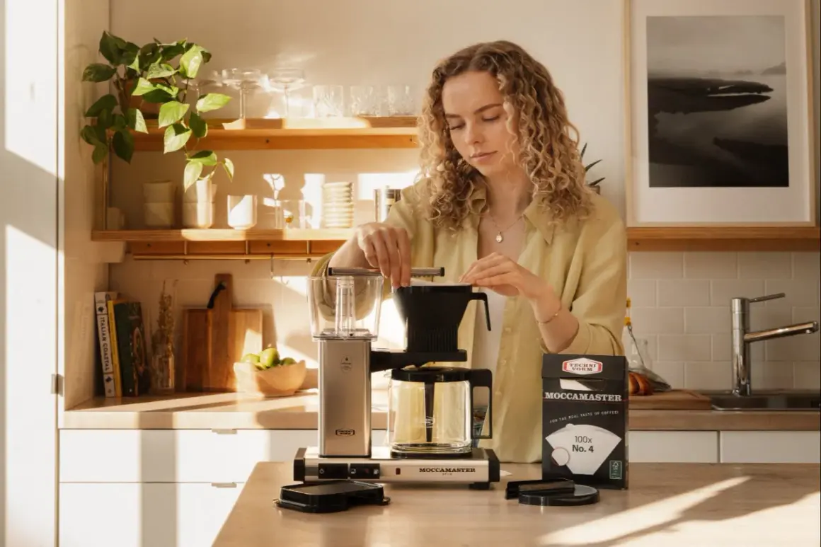 En kvinna förbereder kaffe med en Moccamaster. På bordet ligger en kartong med kaffefilter, där det står "100x No. 4". Det ger en ledtråd om hur många kaffemått per kopp som kan behövas.