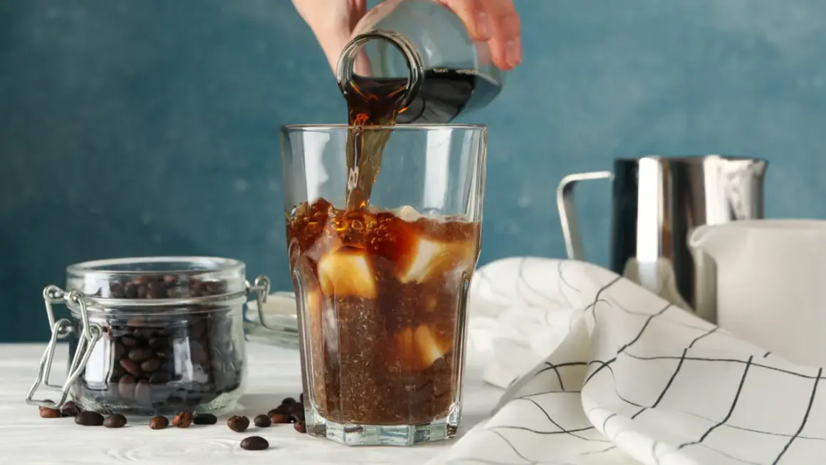 En hand häller mörkt kaffe över is i ett glas. Bredvid står en burk med kaffebönor. Kaffe smakar surt om det inte är perfekt bryggt.