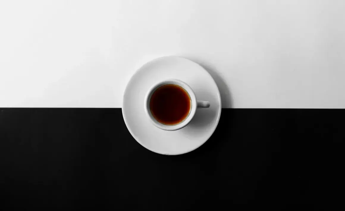 En vit kopp med mörkt kaffe står på en vit tefat, delar en yta som är uppdelad i svart och vitt. Kan kaffe bli gammalt?