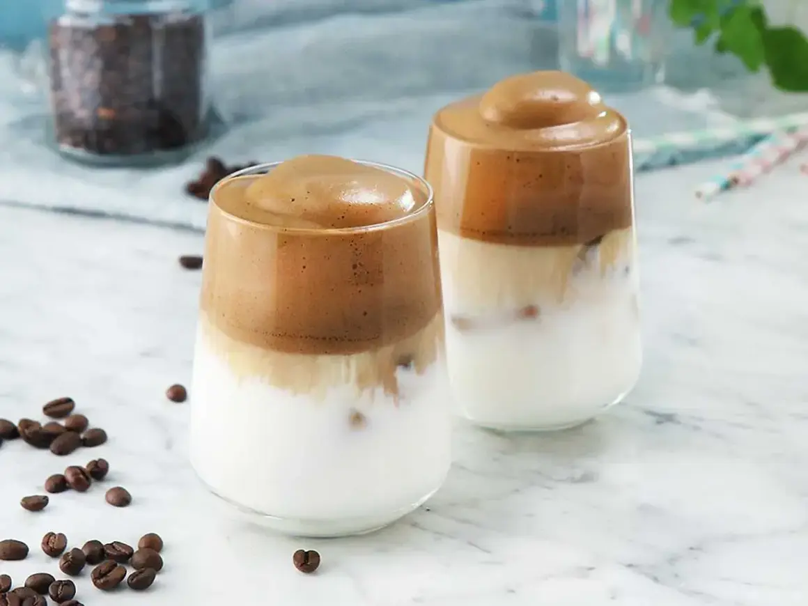 Två glas med krämiga kaffedrinkar med rom, toppade med fluffigt skum. Kaffebönor sprider sig på bordet.