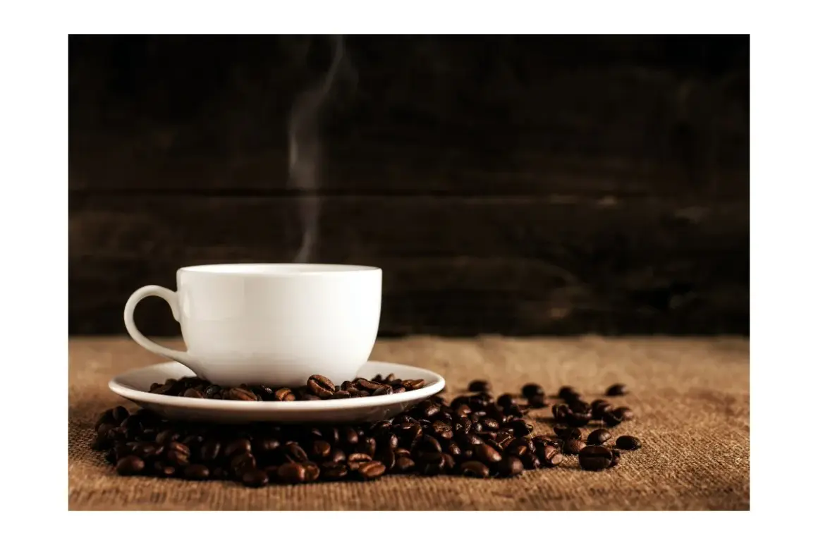 En vit kaffekopp i dl, omgiven av rostade kaffebönor, med ånga som stiger upp.